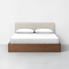 A modern bed with a light beige upholstered headboard, crisp white bedding, and pillows, featuring a brown wooden frame, set against a plain white wall and a light gray floor.