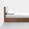 Side view of a minimalist wooden bed with a white mattress, white pillow, and a padded light beige headboard against a plain white wall on a light gray floor.