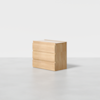 A minimalist light wood dresser with three drawers stands on a plain, light gray surface against a white background. The dresser has clean lines and a modern, simple design.