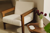 A wooden armchair with cream upholstery sits next to a small wooden table holding a speckled vase of dark red flowers and two white coasters, in a sunlit corner of a room.