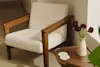 A wooden armchair with cream upholstery sits next to a small wooden table holding a speckled vase of dark red flowers and two white coasters, in a sunlit corner of a room.