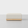 A modern, square-shaped ottoman with a light beige cushion and a light wood base, placed on a smooth, light grey surface against a plain white background.