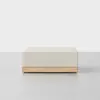 A modern, square-shaped ottoman with a light beige cushion and a light wood base, placed on a smooth, light grey surface against a plain white background.