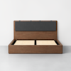 A modern wooden bed frame with a raised slatted base and a dark gray upholstered headboard, set against a plain white wall and a light gray floor.
