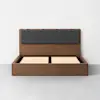 A modern wooden bed frame with a raised slatted base and a dark gray upholstered headboard, set against a plain white wall and a light gray floor.