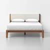 A modern bed with a wooden frame and a light upholstered headboard, neatly made with white bedding and two white pillows, set against a plain white background.