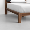 A close-up view of a wooden bed frame with clean lines and a white mattress and pillow, set on a light grey floor against a white wall.