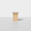 A minimalist wooden bench with a rectangular, light-colored seat and solid base, set against a plain white background.
