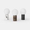 Three modern table lamps with spherical white glass tops and cylindrical marble bases in white, brown, and black, are arranged side by side on a plain white background. Each base has a small circular button.