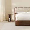 A modern, minimalist wooden nightstand with a flat top and base, positioned next to a wooden bed frame with a white mattress and pillow against a white wall and floor.