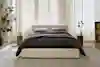 A modern bedroom with a beige upholstered bed frame, dark bedding, two pillows, and matching wooden nightstands with decor. Large windows with white curtains let in natural light. A potted plant is on the left.
