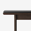 A close-up view of a wooden bench with a dark gray cushioned seat, shown against a white background. Only part of the bench is visible, focusing on the corner and one leg.