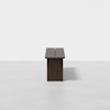 A minimalistic, rectangular wooden bench with clean lines and a dark finish, placed on a light gray floor against a plain white background. The bench is viewed from the end, showing its slim profile.
