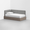 A modern daybed with a light wood base, a white mattress, a white pillow, and gray upholstered back and side panels, positioned in the corner of a minimal, empty room.