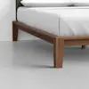 Close-up of a modern wooden bed frame with clean lines and angled legs, supporting a neatly made bed with white bedding, set on a smooth, light gray floor against a plain white wall.