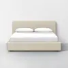 A modern beige upholstered bed with a low headboard, neatly made with white sheets and two white pillows, set against a plain white wall and a light gray floor.