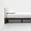 A minimalist bed with a light beige upholstered headboard, white bedding, and a dark wooden frame is positioned against a plain white wall on a light floor.
