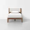 A wooden single bed with a beige upholstered headboard, white bedding, and a single white pillow, set against a plain white wall and light gray floor.