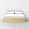A modern bed with a light wood frame, a white upholstered headboard, and white bedding with two pillows, set against a plain white wall and grey floor.