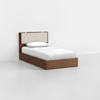 A single bed with a wooden frame and an upholstered beige headboard, made up with white bedding and one pillow, positioned in a minimal, empty white room.