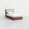 A single bed with a wooden frame and an upholstered beige headboard, made up with white bedding and one pillow, positioned in a minimal, empty white room.