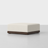 A square, cream-colored ottoman with a textured fabric top and a dark wooden base, placed on a plain light gray floor against a white background.