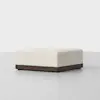 A square, cream-colored ottoman with a textured fabric top and a dark wooden base, placed on a plain light gray floor against a white background.