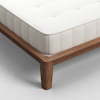 A close-up view of a mattress with a white, quilted surface on a wooden bed frame with sturdy legs, set on a light-colored floor.