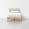 A single bed with a light beige upholstered headboard, white bedding, and a wooden frame, placed against a plain white wall on a light gray floor.