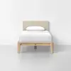 A single bed with a light beige upholstered headboard, white bedding, and a wooden frame, placed against a plain white wall on a light gray floor.