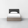 Front view of a modern twin bed with a dark gray upholstered headboard, white bedding, and a light wood frame, set against a plain white wall and light gray floor.