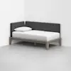 A modern daybed with a light wood frame, white mattress and pillow, and dark gray upholstered back and side panels, placed in the corner of a minimal, white room with a gray floor.