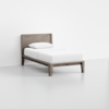 A single bed with a wooden frame and headboard, neatly made with white sheets and a pillow, sits on a light gray floor against a plain white wall.
