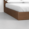 Close-up of the corner of a wooden platform bed with a white mattress and pillows on top, set on a smooth, light gray floor against a white wall.