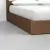 Close-up of the corner of a wooden platform bed with a white mattress and pillows on top, set on a smooth, light gray floor against a white wall.