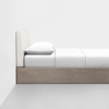 A minimalist bed with a light wood frame, white mattress, white sheets, and a single white pillow, positioned against a plain white wall on a smooth gray floor.