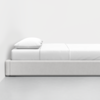 A minimalist bed with a white headboard, white sheets, and a single white pillow, positioned against a plain white wall on a light gray floor.