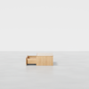 A minimalist wooden bench with a storage drawer partially open, set against a plain white background and placed on a smooth, light grey floor.