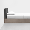 A minimalist bed with a light wood frame, a dark upholstered headboard, and white bedding is positioned against a plain white wall in a bright, uncluttered room.