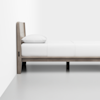 A minimalist wooden bed with a headboard, made up with white bedding and a single pillow, is positioned against a white wall on a light grey floor.