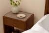 A wooden bedside table with a beige top holds a white round vase with green branches and a white dish containing small jewelry; part of a bed with white bedding is visible beside it.