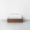 A minimalist single bed with a wooden frame, neatly made with white bedding and a single pillow, set against a plain white wall in a bright, empty room.