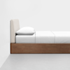A minimalist bed with a light fabric upholstered headboard, white bedding, and a wooden frame, placed against a plain white wall on a gray floor.