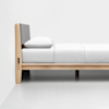 A minimalist wooden bed with a light-colored frame, gray padded headboard, and white bedding, including a pillow and fitted sheet, placed against a plain white wall on a smooth floor.