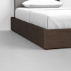 A close-up view of a modern bed with a dark wooden frame, white bedding, and a light grey headboard, set on a light-colored floor against a white wall.
