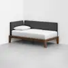 A modern daybed with a wooden frame, white mattress and pillow, and dark gray upholstered back and side panels, placed in the corner of a minimal, white-walled room with a light gray floor.