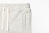 Close-up of the upper part of light gray sweatpants featuring an elastic waistband with cream-colored drawstrings and a side pocket, set against a plain white background.