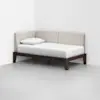 A minimalist daybed with beige upholstered back and sides, a white mattress, a single pillow, and a dark wooden frame, placed in the corner of a white room with a gray floor.