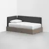 A modern daybed with a light wood base and dark gray upholstered backrests sits in the corner of a minimalist, white room. The bed is made with white bedding and a single pillow.