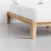 A close-up of a wooden bed frame with a natural finish, supporting a bed with white sheets and pillows, set on a smooth, light gray floor against a white wall.